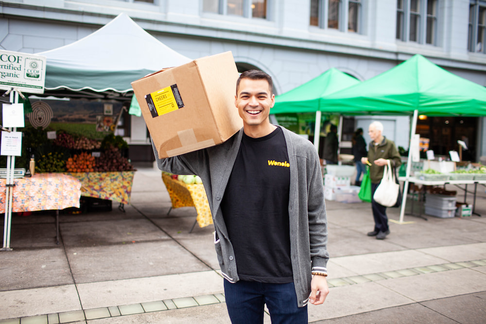 A man is holding a box on his shoulder and smiling in front of his temp job at the farmers market.