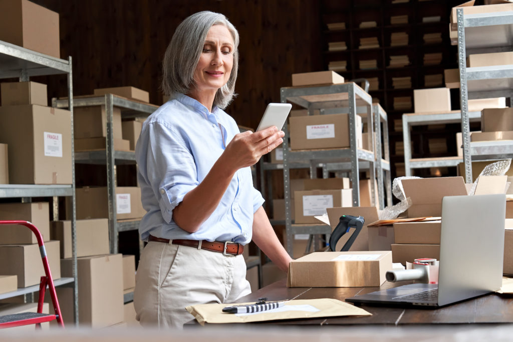 A woman stands at a desk surroundedby boxes and smiles at her phone while choosing her peak season workers.