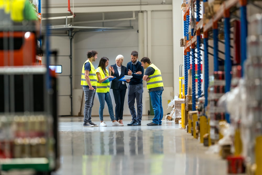 A group or workers in a warehouse stand in a group looking over job responsibilities.